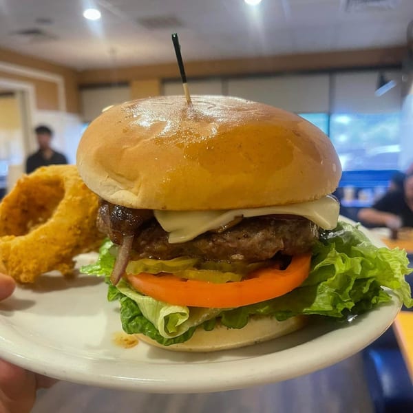 A Good Day Cafe cheeseburger with lettuce, tomato, pickles, and onion rings on a white plate.