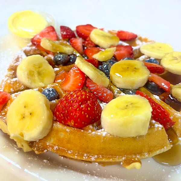 Golden waffle topped with sliced bananas, strawberries, blueberries, powdered sugar, and syrup, with a side of butter.