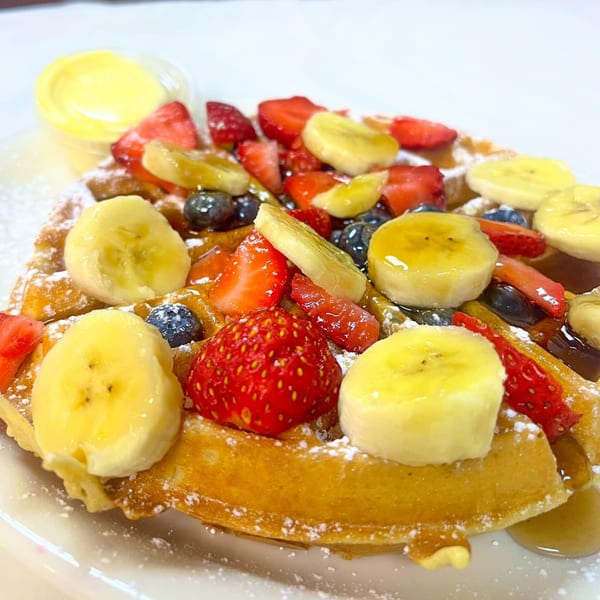 Golden waffle topped with sliced bananas, strawberries, blueberries, powdered sugar, and syrup.