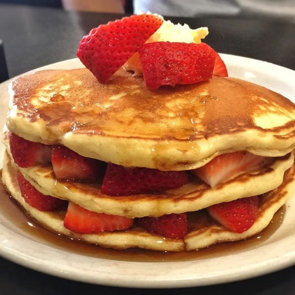Stack of Good Day Cafe pancakes with strawberries and syrup on a white plate.
